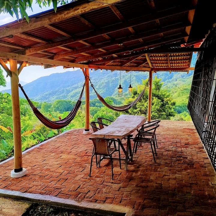 Terraza techada con piso de ladrillo, dos hamacas, mesa larga y el valle verde al fondo.
