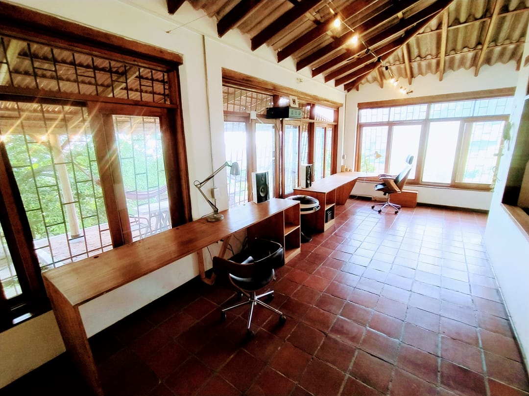 Sala larga con escritorios corridos junto a las ventanas, sillas y piso de baldosa de barro.