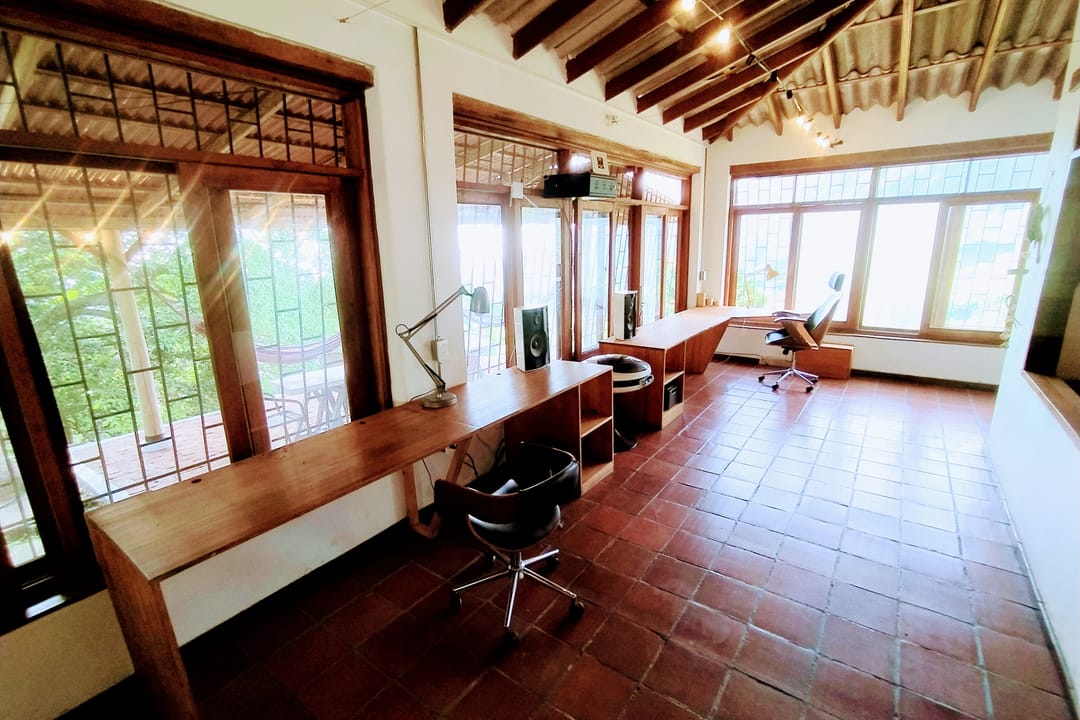 Sala larga con escritorios corridos junto a las ventanas, sillas y piso de baldosa de barro.