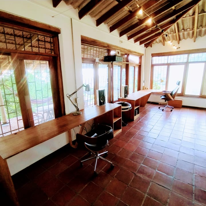 Sala larga con escritorios corridos junto a las ventanas, sillas y piso de baldosa de barro.