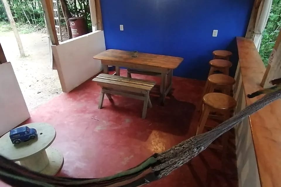 Zona común con pared azul, piso rojo, una hamaca colgada, mesa de madera con banca y butacos altos junto a una baranda hacia la vegetación.