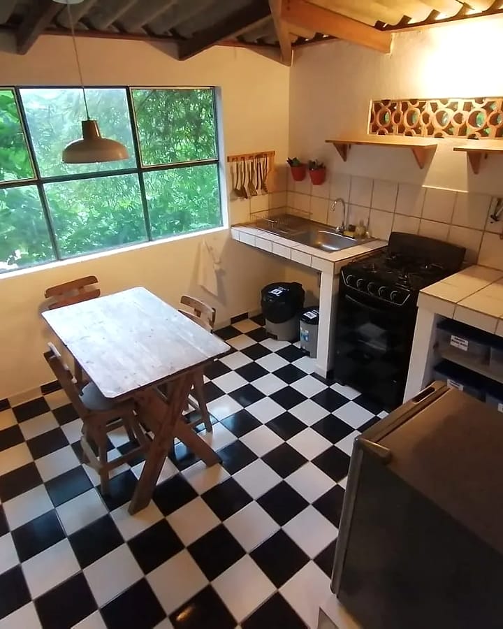 Cocina compartida con piso de ajedrez blanco y negro, estufa a gas, mesones de baldosa, una mesa de madera y una ventana grande hacia el verde.