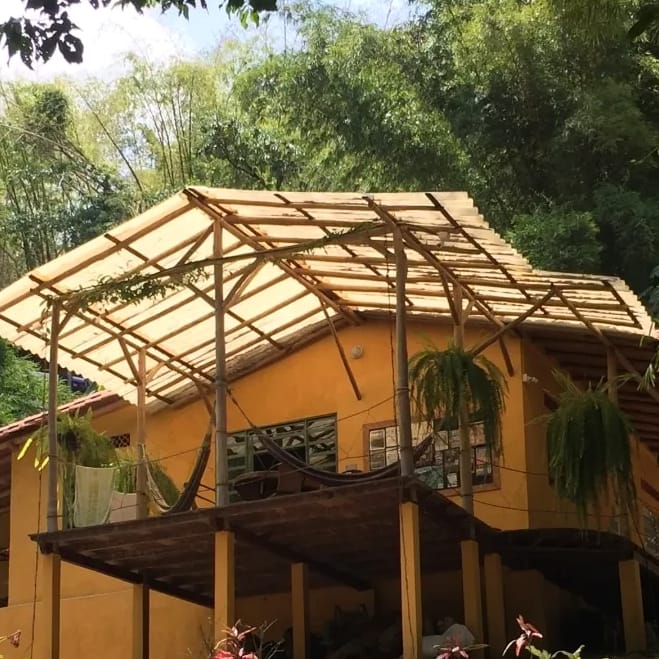 Casa de dos pisos pintada de amarillo, sobre pilotes, con un techo de guadua sobre la terraza alta, plantas colgantes y la selva detrás.