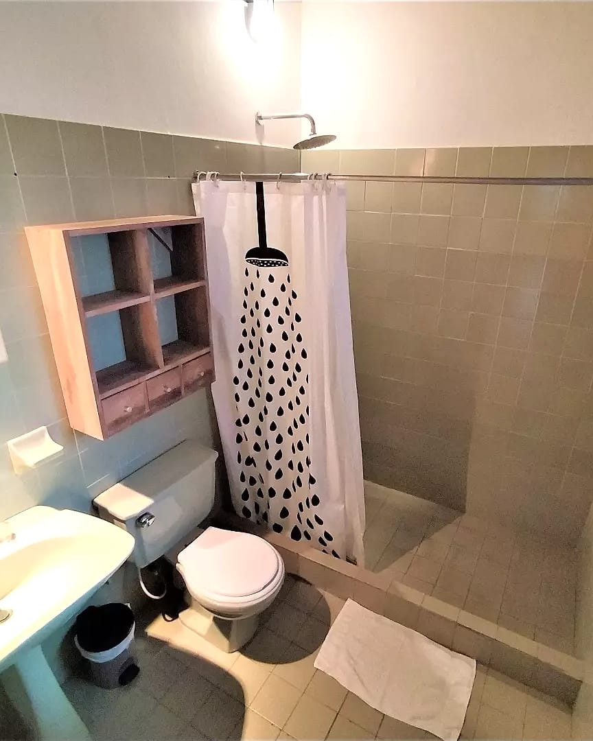 Otro baño de baldosas verde pálido, con ducha de cortina de gotas, inodoro, lavamanos y un mueble de madera.