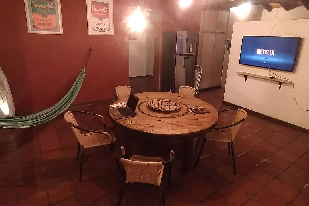 Sala común de pared roja con una mesa redonda de madera, un portátil abierto, una hamaca, lámparas colgantes y un televisor en la pared.