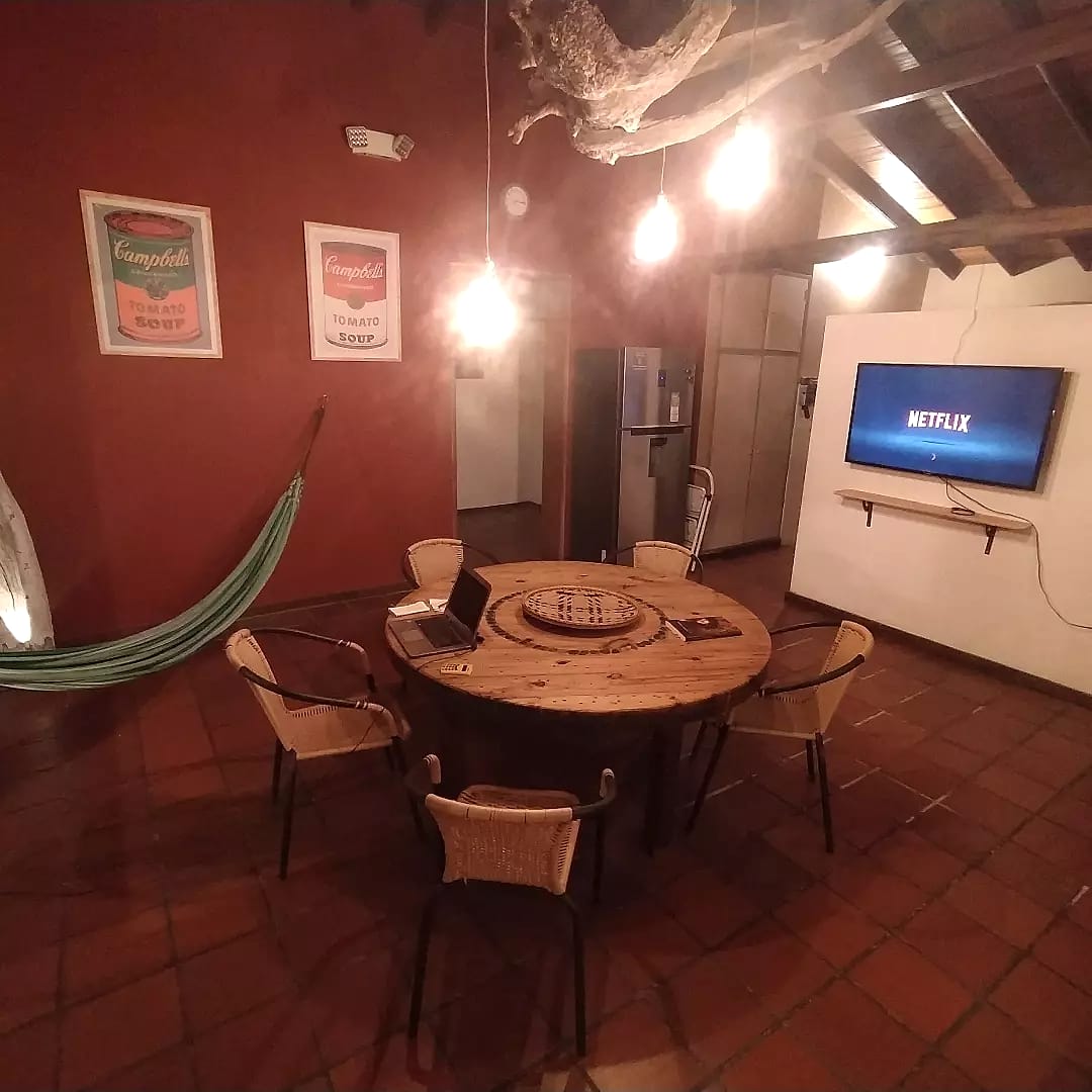 Sala común de pared roja con una mesa redonda de madera, un portátil abierto, una hamaca, lámparas colgantes y un televisor en la pared.