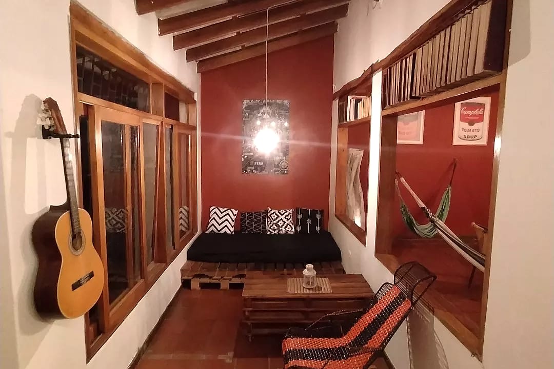 Corredor de descanso con una guitarra colgada, un sofá bajo con cojines, una mecedora tejida, una hamaca y estantes con libros sobre las ventanas.
