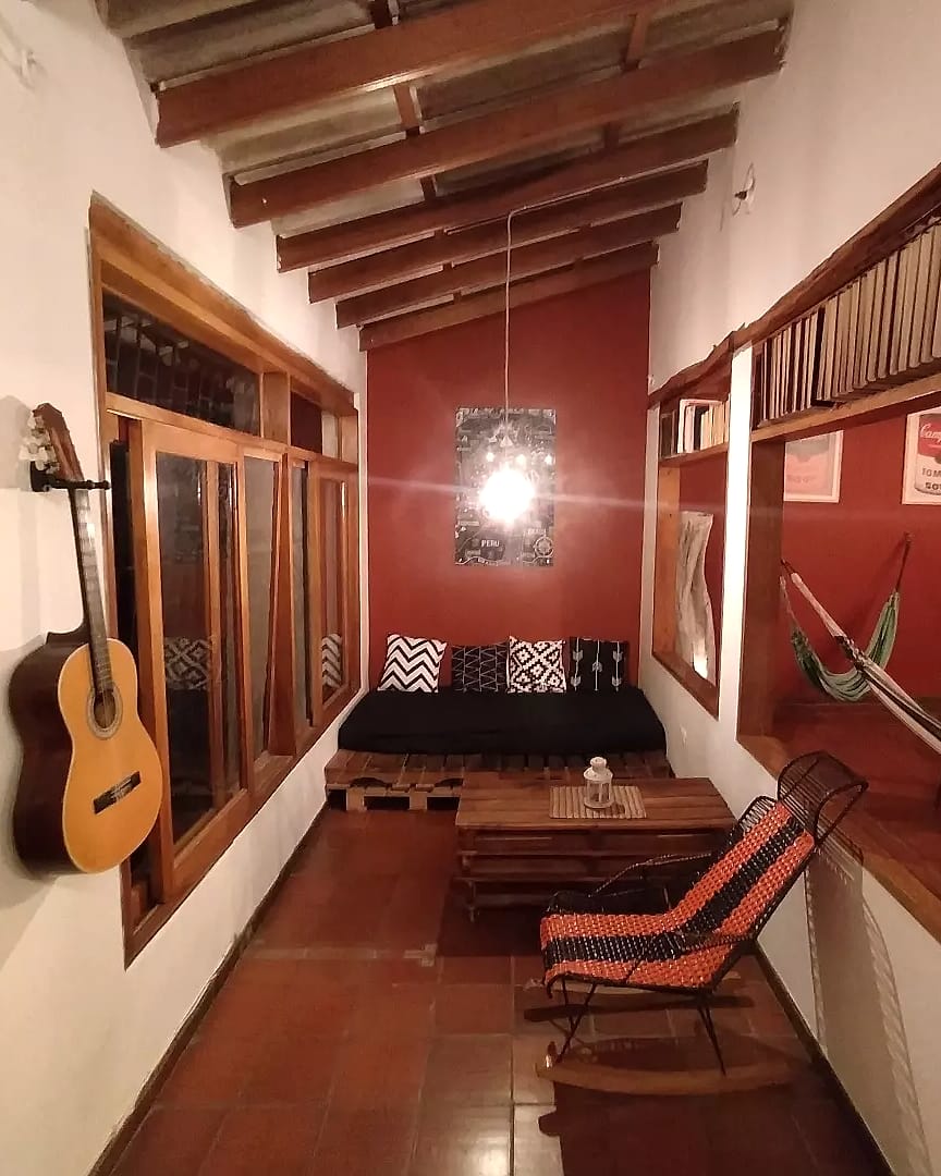 Corredor de descanso con una guitarra colgada, un sofá bajo con cojines, una mecedora tejida, una hamaca y estantes con libros sobre las ventanas.