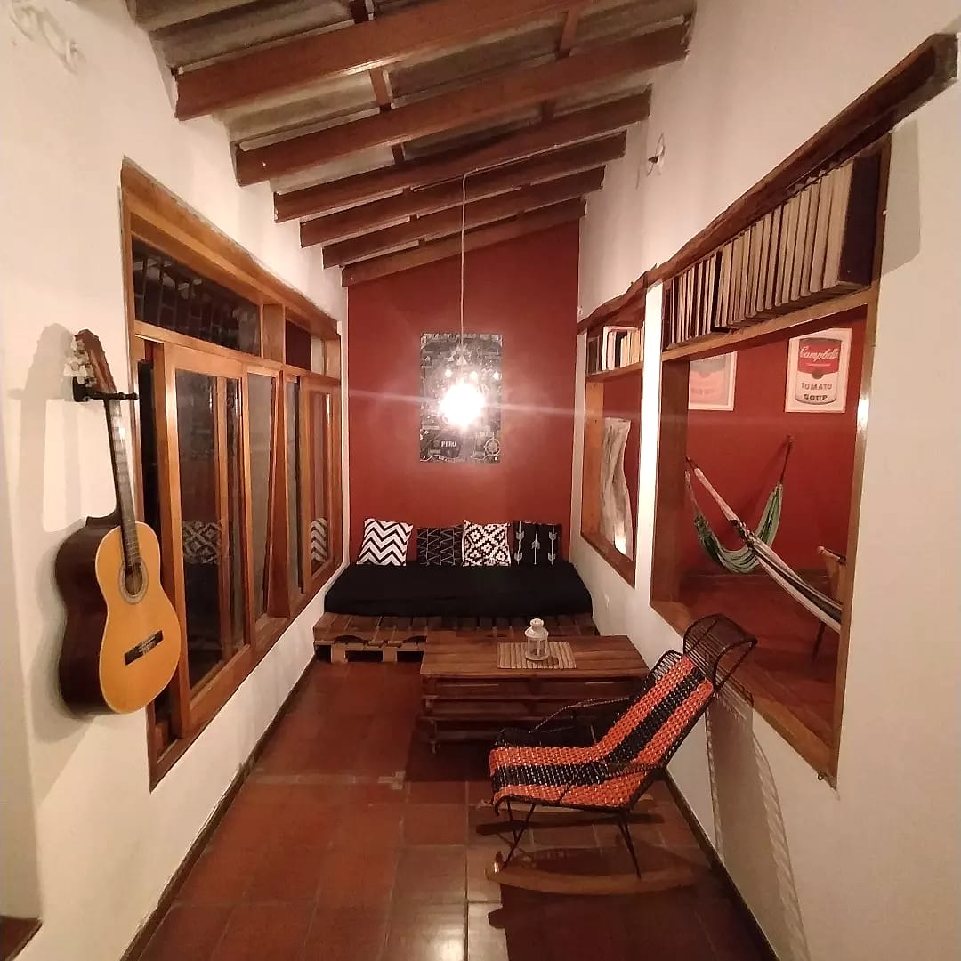 Corredor de descanso con una guitarra colgada, un sofá bajo con cojines, una mecedora tejida, una hamaca y estantes con libros sobre las ventanas.