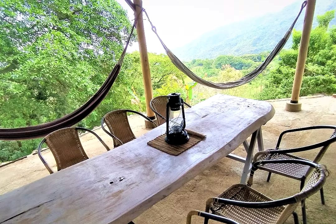 Terraza techada con una mesa larga de madera rústica, un farol, sillas tejidas y dos hamacas; al fondo, el valle verde y las montañas.