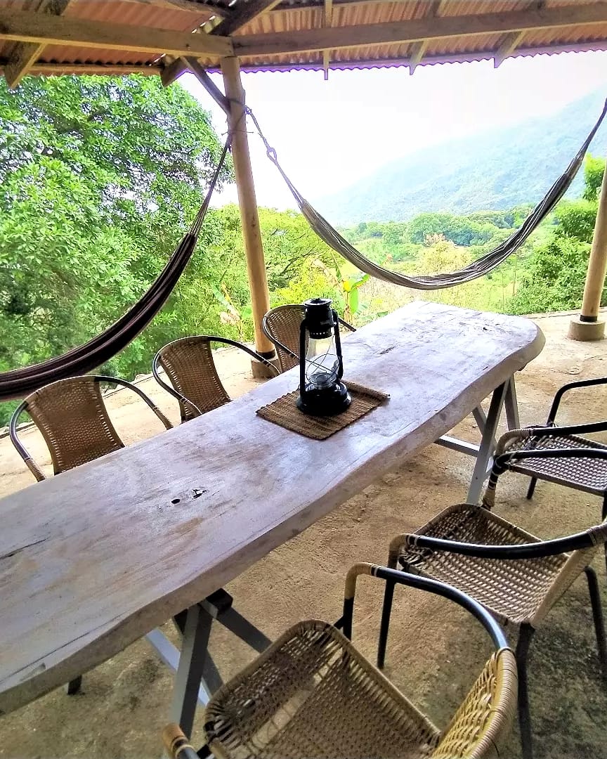 Terraza techada con una mesa larga de madera rústica, un farol, sillas tejidas y dos hamacas; al fondo, el valle verde y las montañas.
