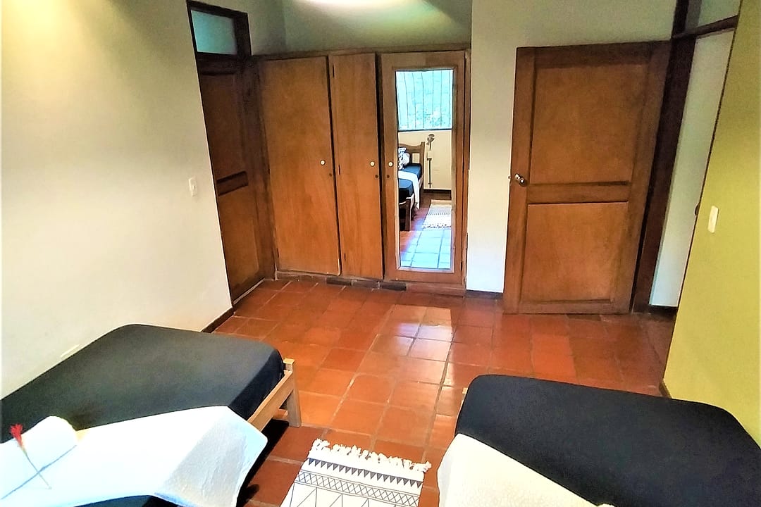 Dos camas sencillas vistas hacia la puerta de madera, con piso de baldosa y un tapete tejido.