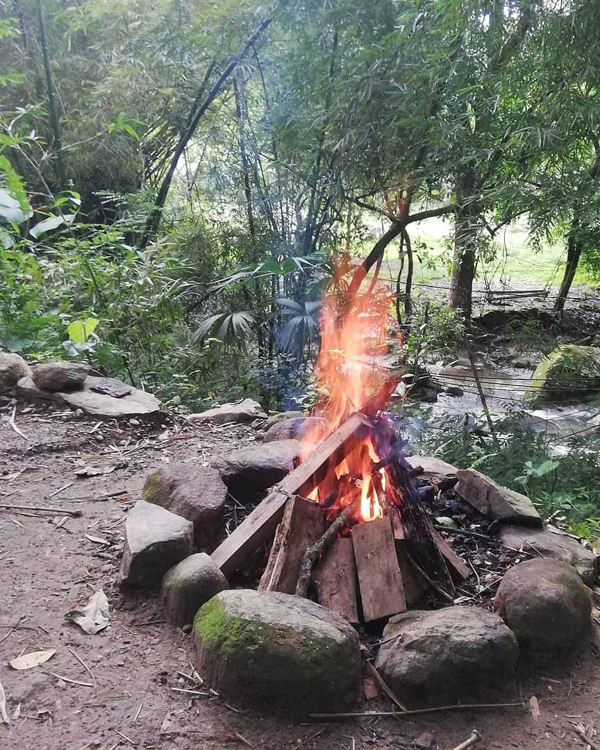 Una fogata en un círculo de piedras junto al río, bajo árboles altos, con las rocas del cauce al fondo.