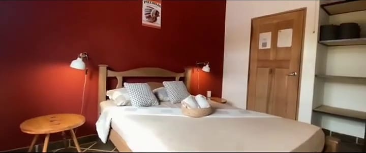 Habitación con cama doble de sábanas blancas, pared roja, lámparas de lectura a cada lado y una puerta de madera.