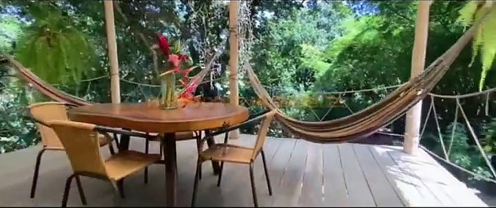 Terraza de madera rodeada de árboles, con hamacas tendidas, una mesa redonda con flores de heliconia y sillas.