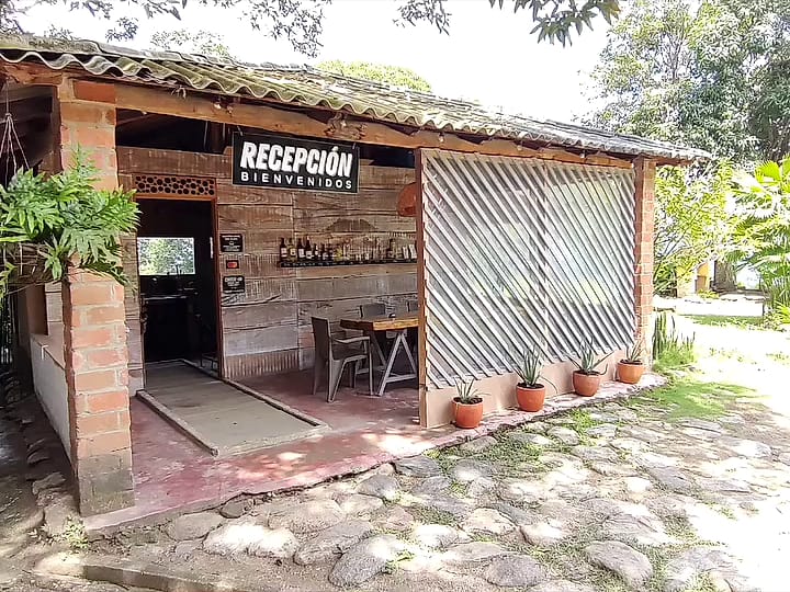 Construcción abierta de madera con el letrero “Recepción · Bienvenidos”, piso de piedra y matas en macetas de barro.