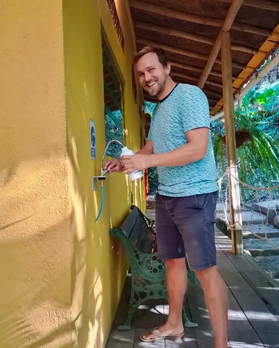 Un huésped llena una botella reutilizable en el dispensador de agua filtrada, junto a una pared amarilla y un corredor de madera.
