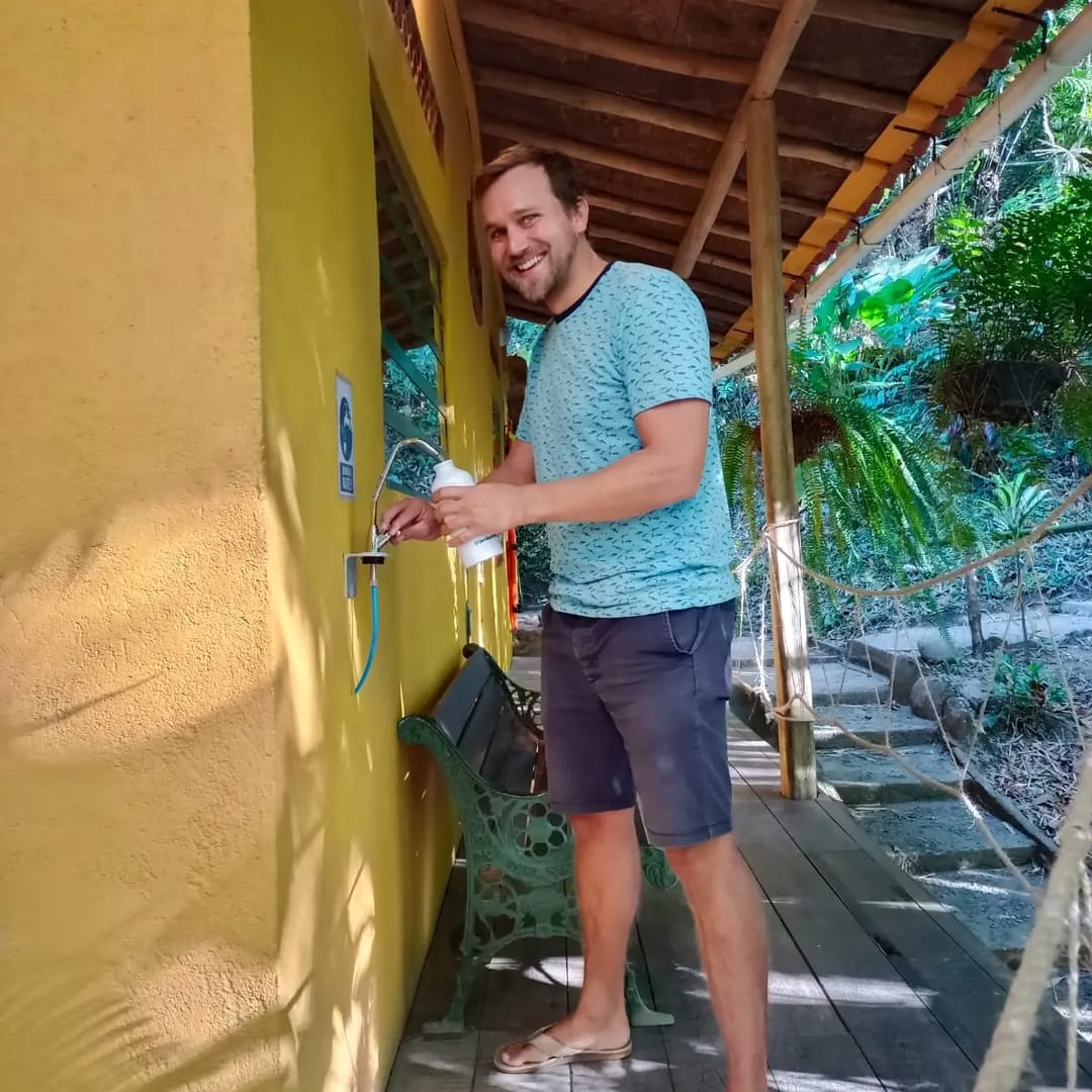 Un huésped llena una botella reutilizable en el dispensador de agua filtrada, junto a una pared amarilla y un corredor de madera.